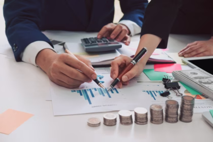 Dois profissionais analisando gráficos financeiros em uma mesa com calculadora, moedas empilhadas e notas, representando a análise de custo de desenvolvimento de software