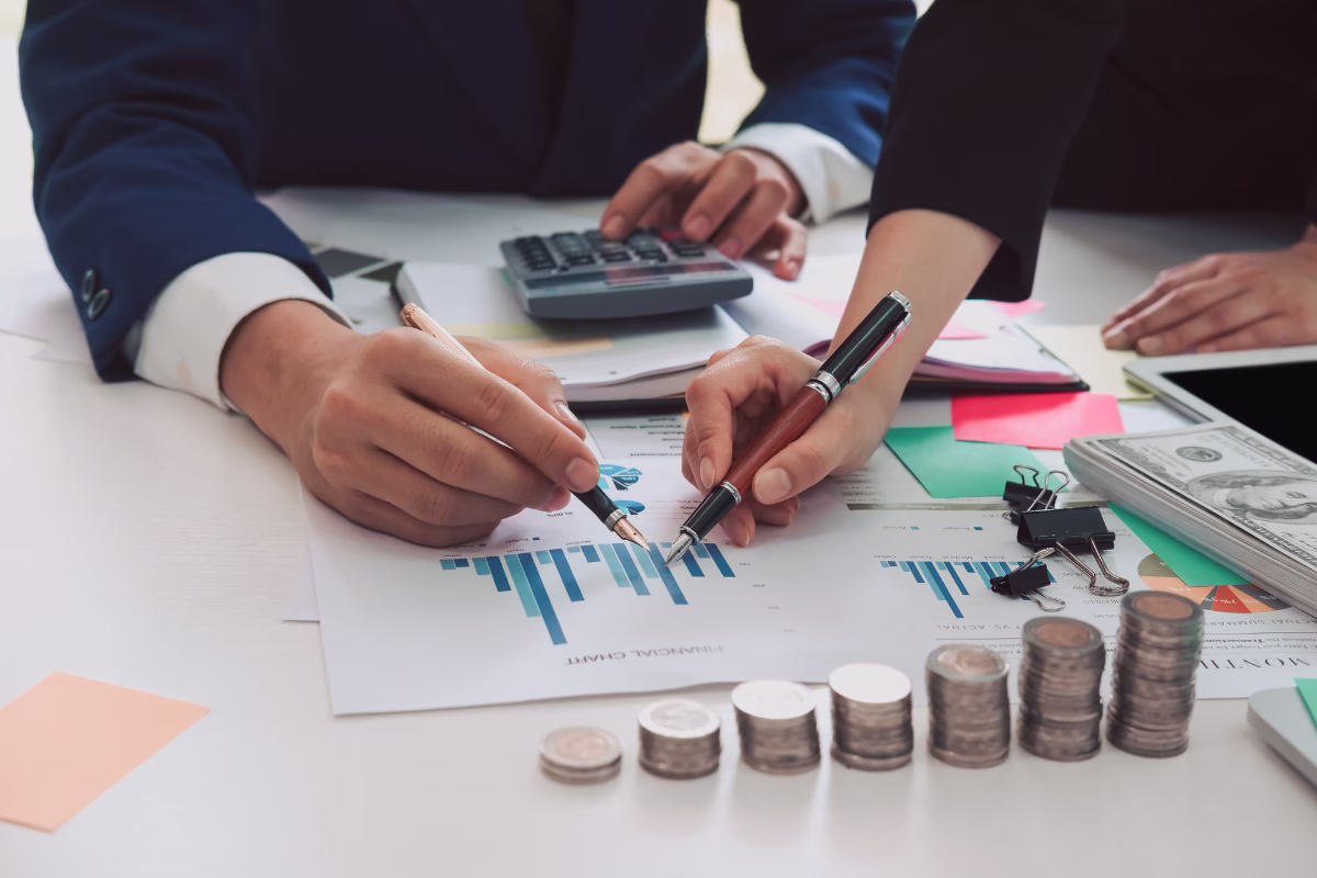Dois profissionais analisando gráficos financeiros em uma mesa com calculadora, moedas empilhadas e notas, representando a análise de custo de desenvolvimento de software
