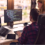 Desenvolvedor e colega revisando código TypeScript em monitor de escritório