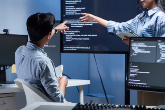 Profissionais de tecnologia analisando código de um modelo de inteligência artificial em monitores durante reunião de equipe de desenvolvimento de software