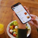 Mão segurando um smartphone exibindo o cardápio digital do Restaurante VINA, com opções de pizzas e pratos quentes, ao lado de uma salada e um copo de suco de laranja sobre uma mesa de madeira.