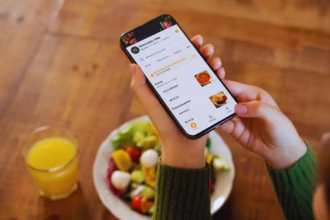 Mão segurando um smartphone exibindo o cardápio digital do Restaurante VINA, com opções de pizzas e pratos quentes, ao lado de uma salada e um copo de suco de laranja sobre uma mesa de madeira.