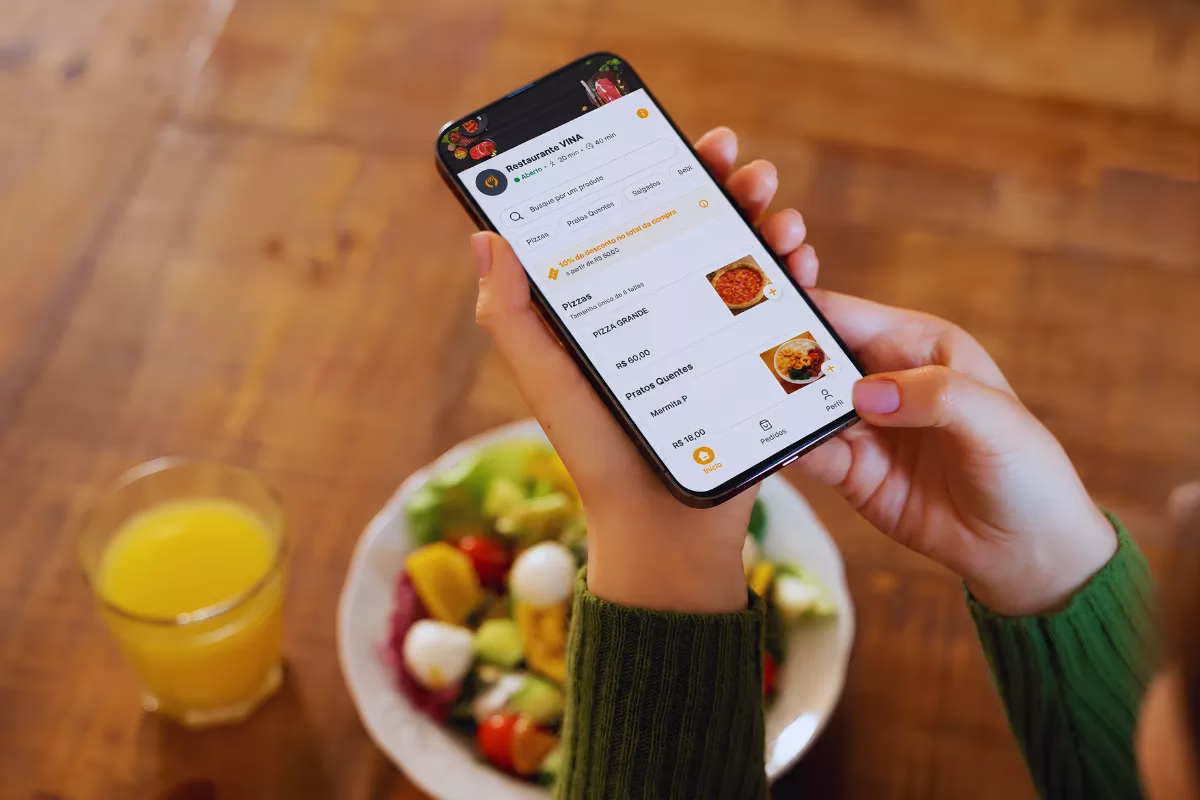 Mão segurando um smartphone exibindo o cardápio digital do Restaurante VINA, com opções de pizzas e pratos quentes, ao lado de uma salada e um copo de suco de laranja sobre uma mesa de madeira.