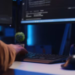 Desenvolvedor digitando em um teclado em frente a monitores com código-fonte, em um ambiente de trabalho com iluminação azul