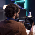 Desenvolvedor revisando código-fonte em um tablet, com monitores exibindo um editor de código ao fundo.