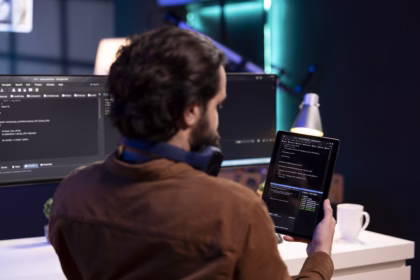 Desenvolvedor revisando código-fonte em um tablet, com monitores exibindo um editor de código ao fundo.