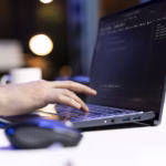 Mão de uma pessoa digitando em um notebook com código exibido na tela, representando o trabalho de desenvolvimento de software.