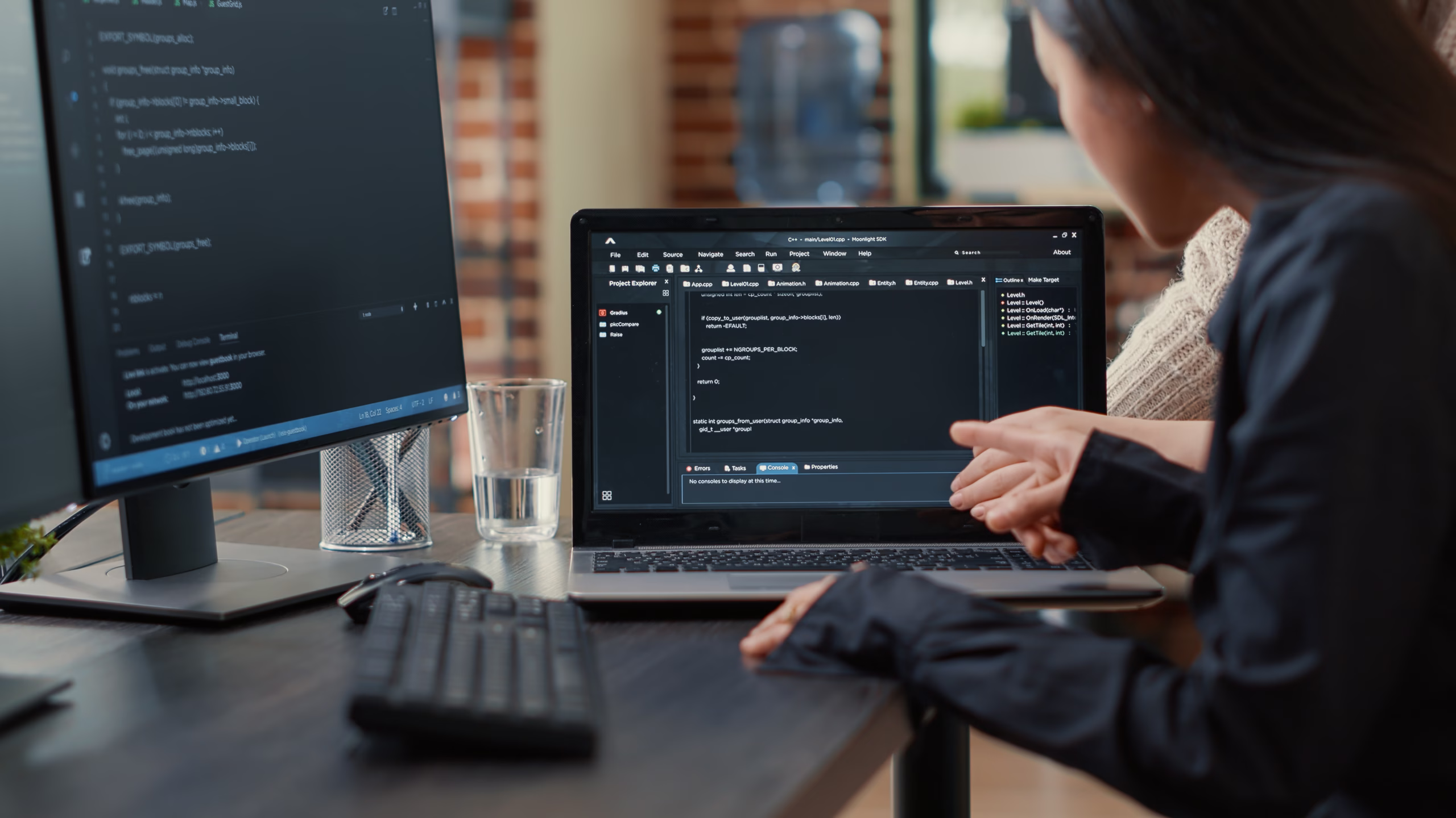 Two developers review source code on a laptop, one pointing at the screen, with a monitor displaying more code beside them.