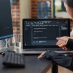 Duas desenvolvedoras analisam código-fonte em um notebook, uma delas apontando para a tela, com um monitor exibindo mais código ao lado.