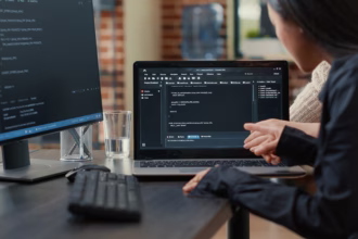 Duas desenvolvedoras analisam código-fonte em um notebook, uma delas apontando para a tela, com um monitor exibindo mais código ao lado.