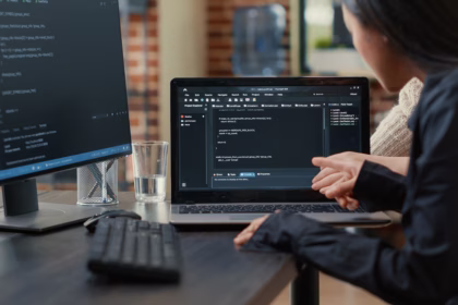 Duas desenvolvedoras analisam código-fonte em um notebook, uma delas apontando para a tela, com um monitor exibindo mais código ao lado.
