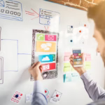 Profissional planeja a interface de um aplicativo em um quadro branco com wireframes, ícones de papel e post-its, segurando um smartphone