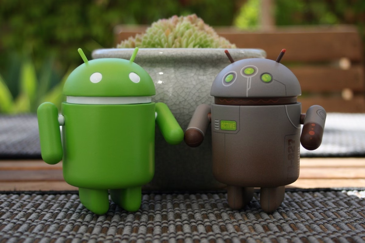 Dois bonecos do robô Android, um verde e um cinza com detalhes metálicos, lado a lado sobre uma mesa, representando o desenvolvimento Android com Kotlin.