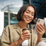 Mulher sorrindo ao olhar para o celular, com um copo de café na outra mão, sentada em uma escada em frente a um prédio corporativo.