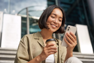 Mulher sorrindo ao olhar para o celular, com um copo de café na outra mão, sentada em uma escada em frente a um prédio corporativo.