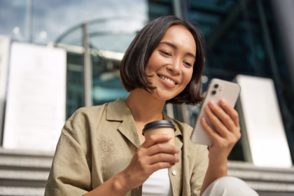 Mulher sorrindo ao olhar para o celular, com um copo de café na outra mão, sentada em uma escada em frente a um prédio corporativo.