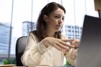 Executiva em escritório com janelas amplas, olhando para o notebook e gesticulando enquanto discute uma decisão de tecnologia