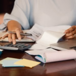 Profissional usando uma calculadora sobre a mesa, com notas fiscais, caderno, post-its e laptop, calculando o custo de contratar uma fábrica de software.