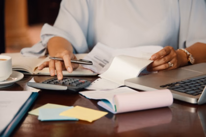 Profissional usando uma calculadora sobre a mesa, com notas fiscais, caderno, post-its e laptop, calculando o custo de contratar uma fábrica de software.