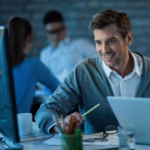 Gestor de TI sorrindo ao trabalhar em um computador em um escritório, com colegas de equipe ao fundo