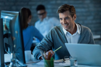 Gestor de TI sorrindo ao trabalhar em um computador em um escritório, com colegas de equipe ao fundo