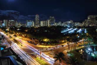 Vista noturna do alto de um cruzamento de avenidas com rastros de luz dos carros em longa exposição, palmeiras e prédios iluminados ao fundo.