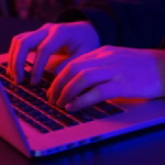 Mãos digitando em um notebook em ambiente escuro, com iluminação em tons de vermelho e roxo, ilustrando a programação de um site.