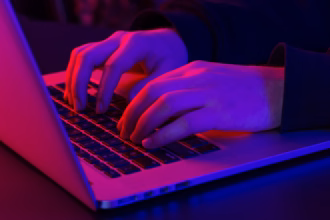 Mãos digitando em um notebook em ambiente escuro, com iluminação em tons de vermelho e roxo, ilustrando a programação de um site.