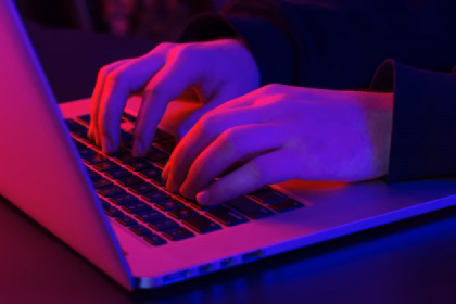Mãos digitando em um notebook em ambiente escuro, com iluminação em tons de vermelho e roxo, ilustrando a programação de um site.