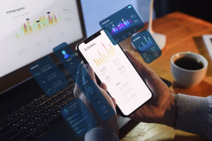 Mão segurando um smartphone com gráfico de linha na tela, cercado por painéis de interface translúcidos (calendário, tela de login, portfólio e assinatura) em tons de azul e rosa, com notebook e caderno desfocados ao fundo.
