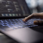 Mão sobre o teclado iluminado de um notebook, com uma tela ao fundo exibindo um fluxo de trabalho com blocos e conexões, representando desenvolvimento de sistemas.