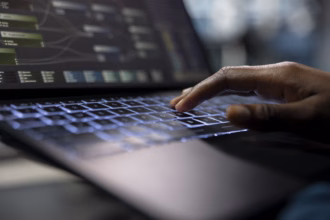 Mão sobre o teclado iluminado de um notebook, com uma tela ao fundo exibindo um fluxo de trabalho com blocos e conexões, representando desenvolvimento de sistemas.