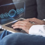 Mãos de uma pessoa digitando em um notebook, com uma interface holográfica translúcida sobre a tela exibindo ícones de escudo com verificação, rede global, equipe, documento e engrenagem, que representam segurança e disponibilidade de sistemas.