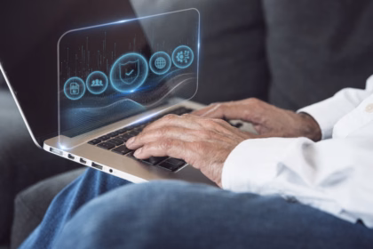 Mãos de uma pessoa digitando em um notebook, com uma interface holográfica translúcida sobre a tela exibindo ícones de escudo com verificação, rede global, equipe, documento e engrenagem, que representam segurança e disponibilidade de sistemas.
