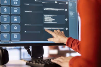 Pessoa de suéter vermelho aponta para a tela de um monitor que exibe uma interface de assistente de IA, com um chat de perguntas e respostas e atalhos para criar vídeo, imagem, calendário e texto.